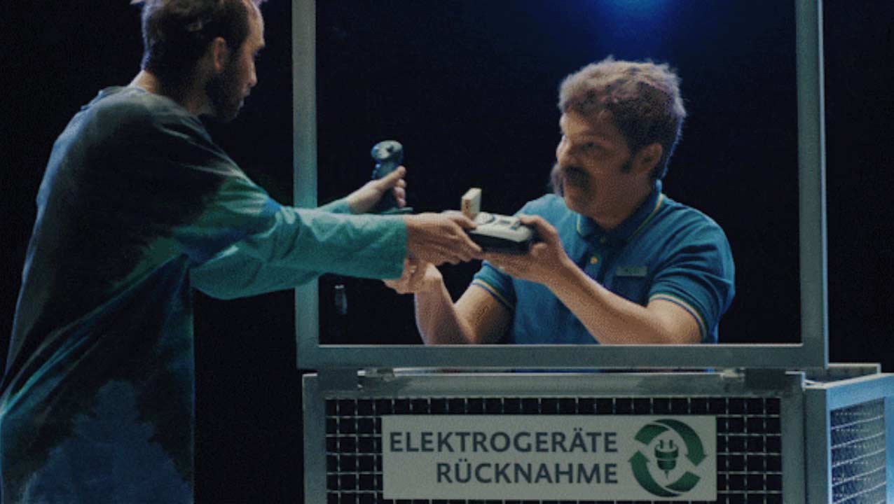 Ein Mann, hinter einem Schild mit dem Text "Elektrogeräte Rückname", nimmt alte Elektrogeräte von einer Person entgegen.
